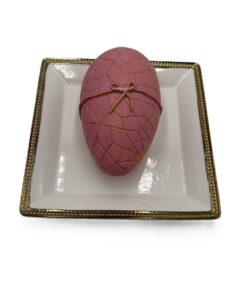 Pink egg-shaped dessert with a brown ribbon on a white plate with a gold-trim edge, decorative and edible-looking.