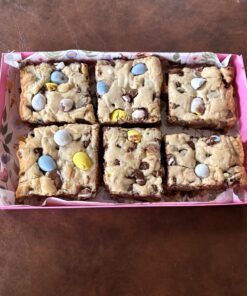 Six square blondie bar desserts with pastel candy eggs in a pink gift box lined with floral paper.