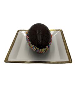 Dark chocolate Easter egg with a geometric pattern rests on a white plate with a gold rim, surrounded by colorful sprinkles at the base.