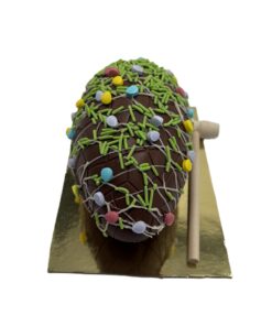 Chocolate egg cake covered in green sprinkles, colorful candy confetti, and white drizzle, on a gold base with a wooden mallet beside it.