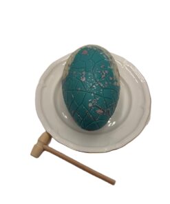 Decorative teal Easter egg with a geometric crack pattern on a white plate, with a small wooden mallet beside it.